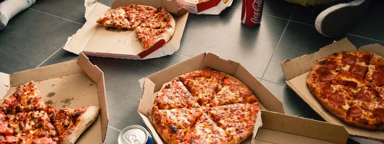 geopende pizzadozen met verse pizza's en cola blikjes eromheen met zittende mensen op de vloer