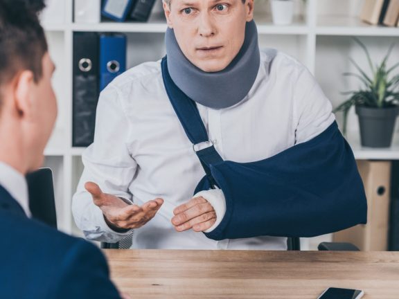 middle aged worker in neck brace with broken arm sitting at table and talking to opposite businessman in office, compensation concept