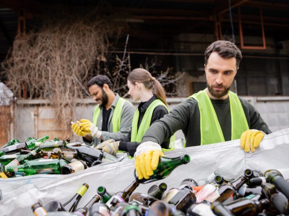 Bearded worker in protective gloves and vest putting glass bottle in sack near blurred interracial colleagues in outdoor waste disposal station, garbage sorting and recycling concept