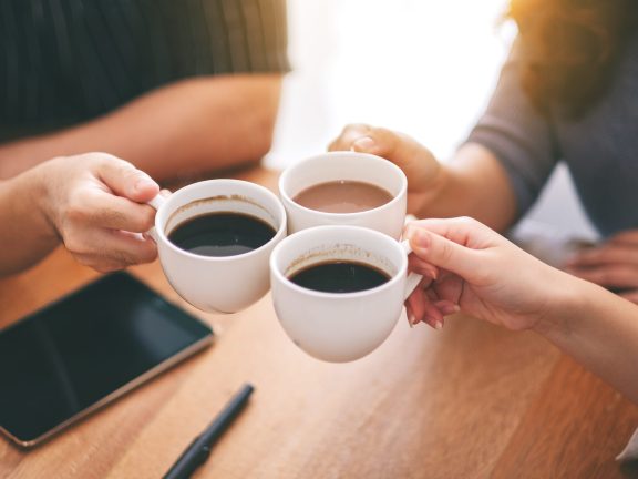 Top view image of three people clinking coffee cups on wooden table in cafe