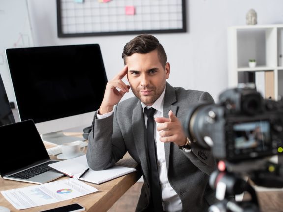 businessman near laptop and computer monitors pointing with finger at digital camera on blurred foreground