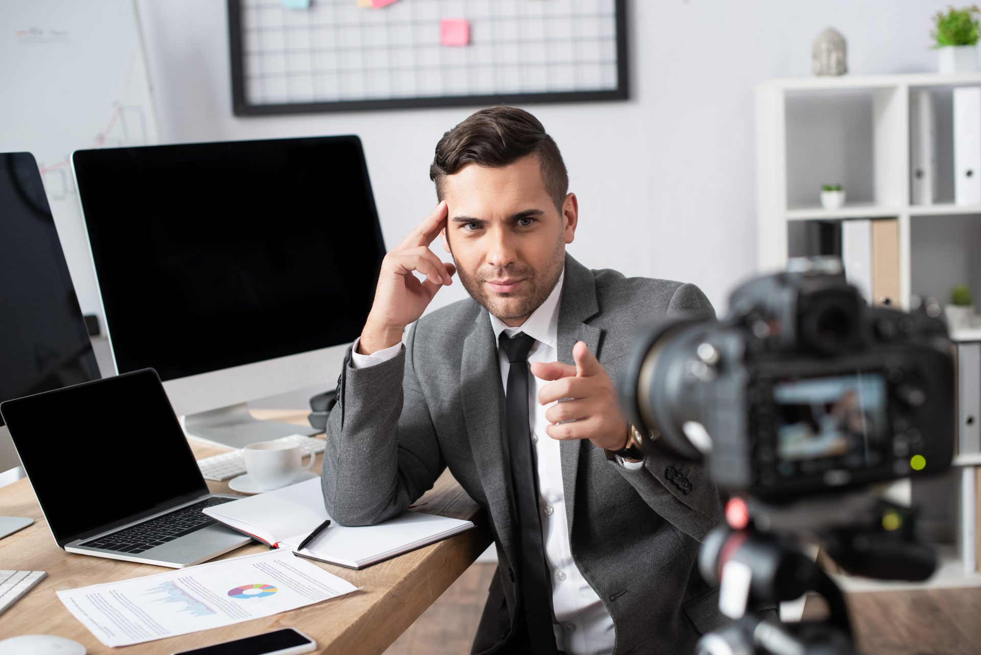 businessman near laptop and computer monitors pointing with finger at digital camera on blurred foreground