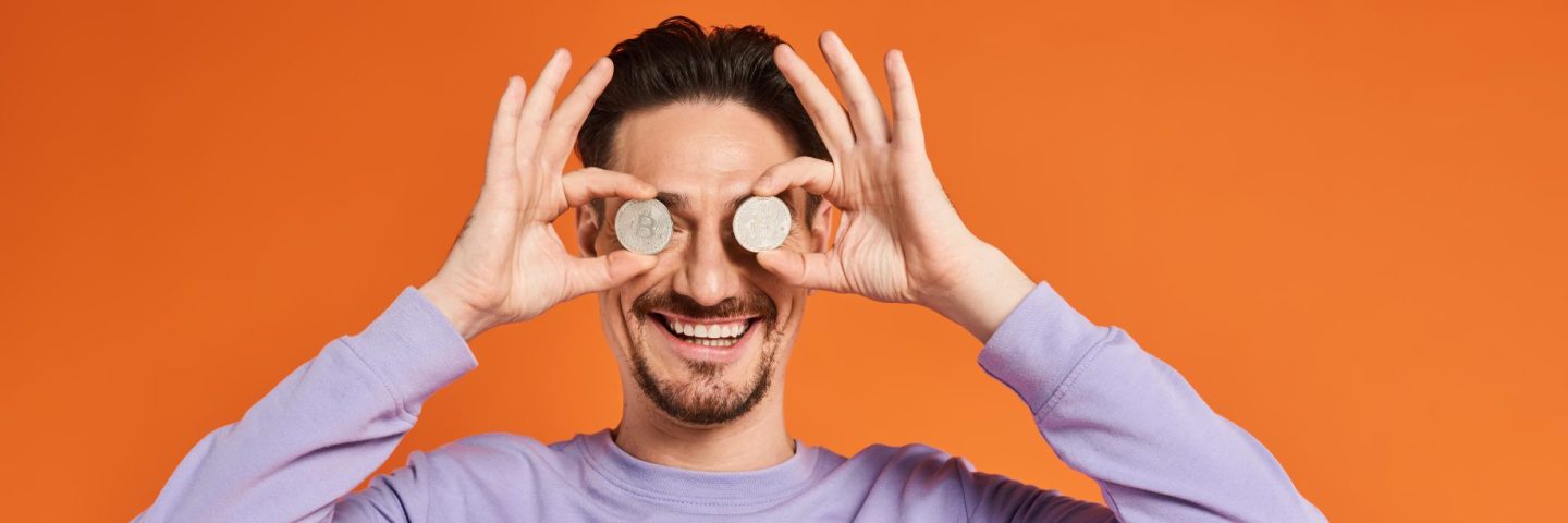 happy man holding bitcoins near eyes and smiling on orange background, cryptocurrency banner