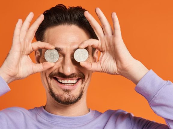 happy man holding bitcoins near eyes and smiling on orange background, cryptocurrency banner