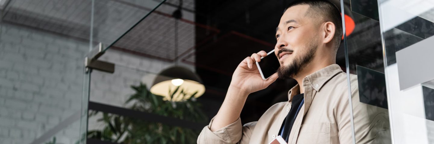 happy asian businessman with tattoo holding folder and talking on smartphone near glass door