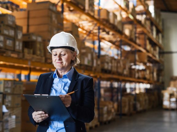 Senior woman warehouse manager or supervisor with white helmet and clipboard.