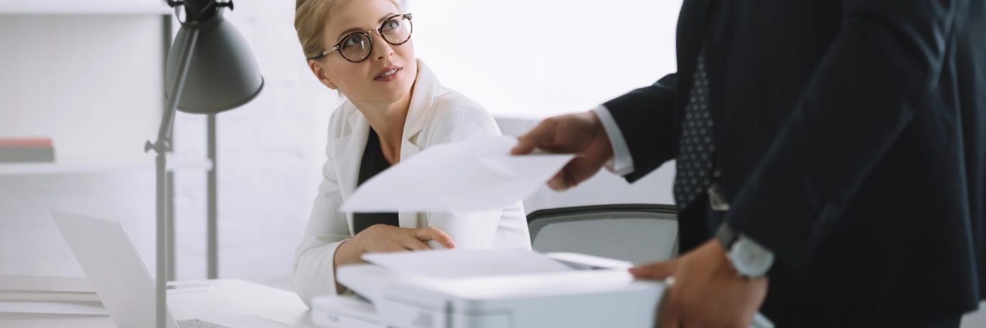 selective focus of businesswoman in eyeglasses looking at colleague with papers near by in office