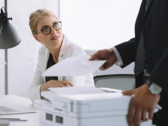 selective focus of businesswoman in eyeglasses looking at colleague with papers near by in office