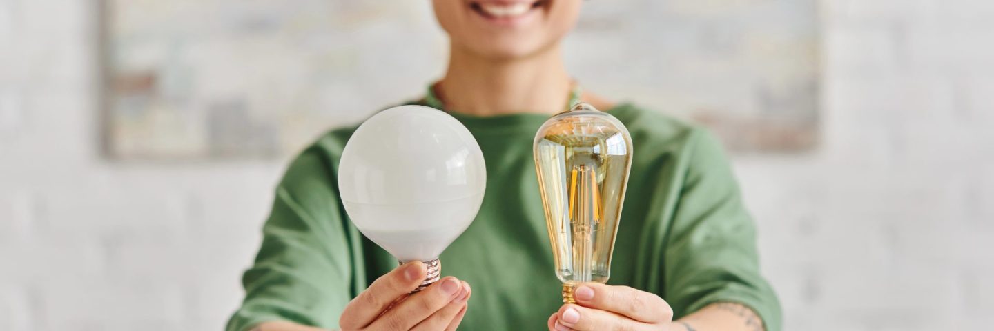 cheerful, young and tattooed woman in casual clothes showing energy saving light bulbs and looking at camera, environmentally conscious, sustainable lifestyle and environmentally conscious concept