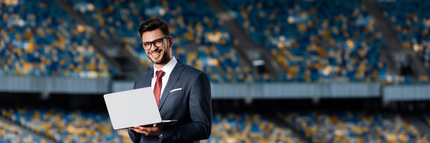 smiling young businessman in suit with laptop at stadium