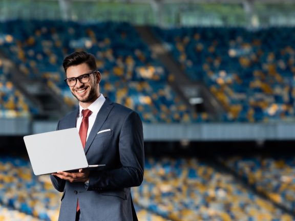 smiling young businessman in suit with laptop at stadium