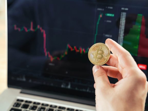 Closeup image of male hands holding golden bitcoin on background with laptop screen with financial chart data, crypto trader checking and controlling financial database on international exchanges