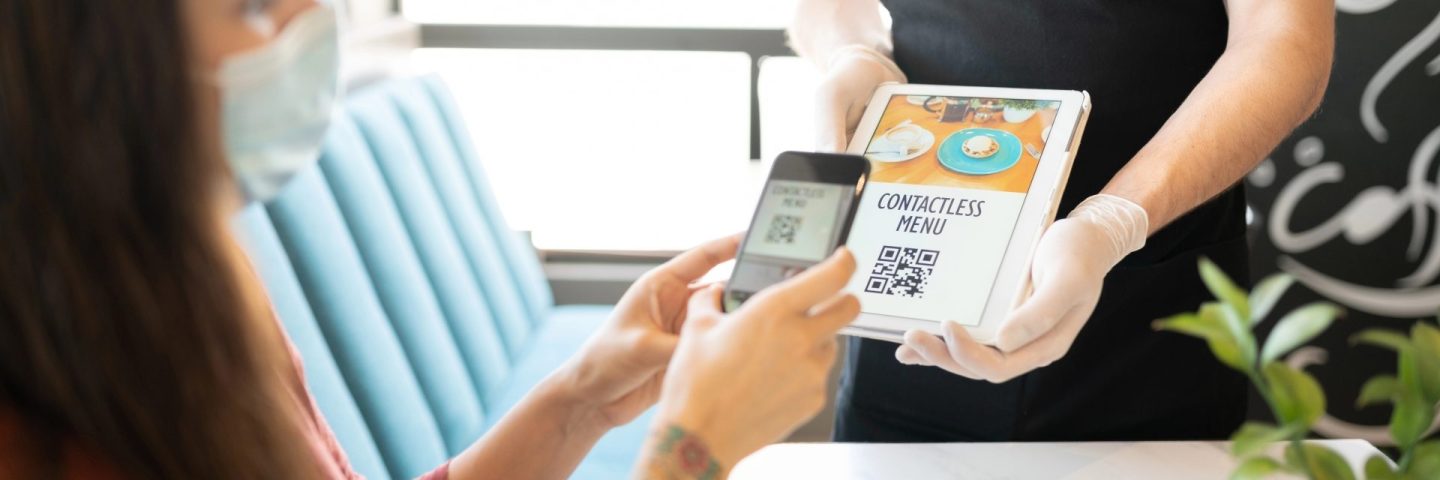 Young woman scanning QR code of contactless menu through smartphone in restaurant