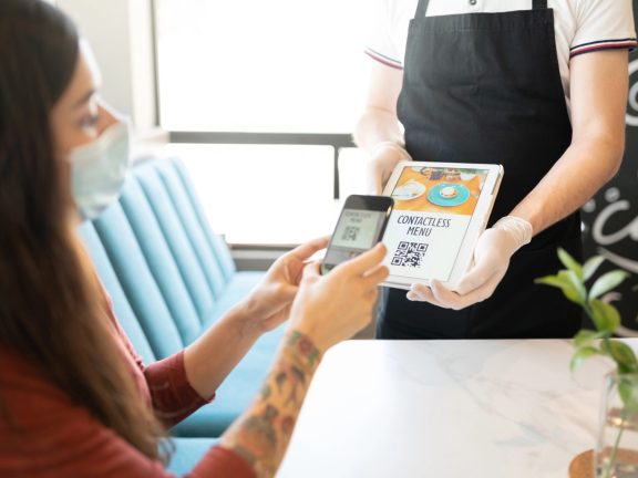 Young woman scanning QR code of contactless menu through smartphone in restaurant