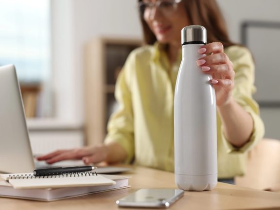Woman holding thermos bottle at workplace, focus on container. Space for text