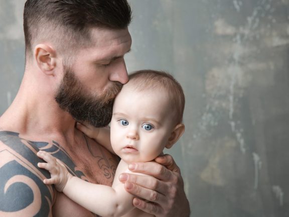 Handsome tattooed young man holding cute little baby on gray background