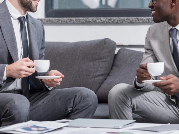 successful multiethnic businessmen sitting on couch and drinking coffee in office