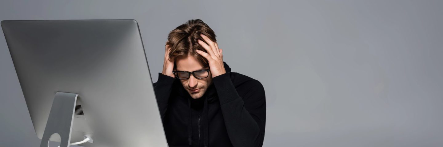 Upset programmer sitting near computer and cellphone on table isolated on grey