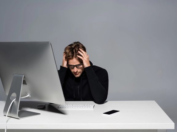 Upset programmer sitting near computer and cellphone on table isolated on grey