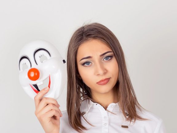 Doubtful inside. Wearing a mask. Portrait young doubtful worried woman with skeptical expression holding a clown mask expressing cheerfulness happiness looking at camera isolated white background.