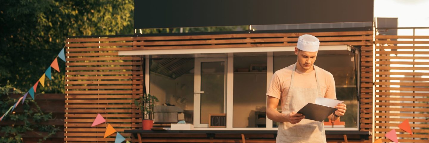 handsome chef in apron looking at clipboard near food truck