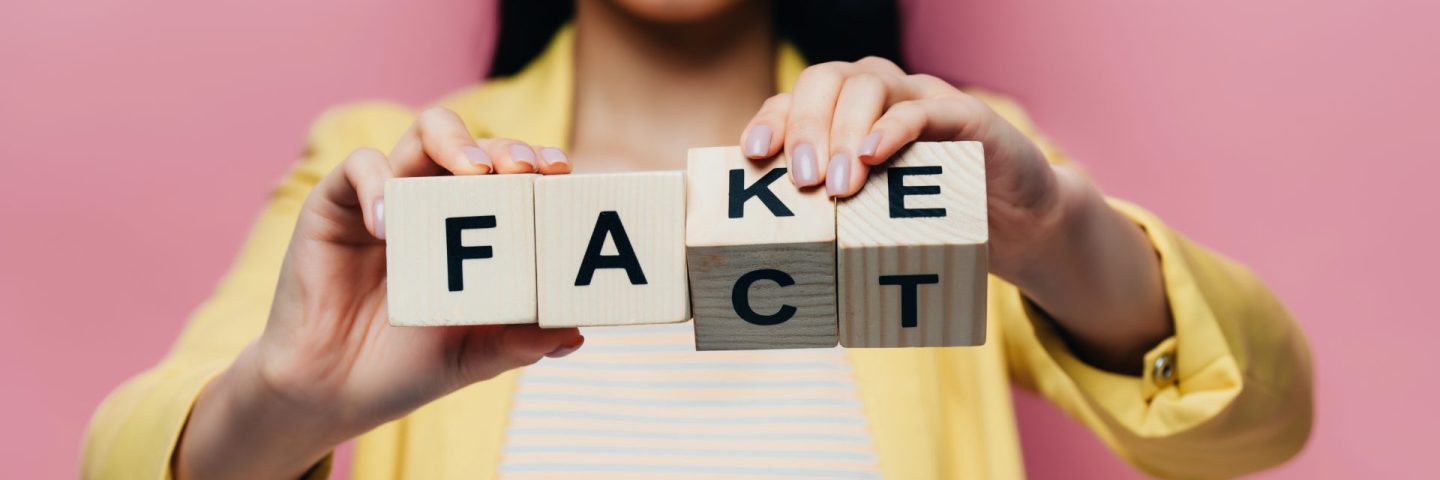 cropped view of asian woman holding wooden cubes with fake and fact lettering isolated on pink