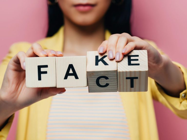 cropped view of asian woman holding wooden cubes with fake and fact lettering isolated on pink