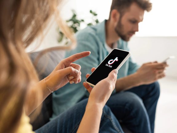 KYIV, UKRAINE - FEBRUARY 21, 2020: Selective focus of woman pointing with finger at smartphone with TikTok app near boyfriend chatting on couch