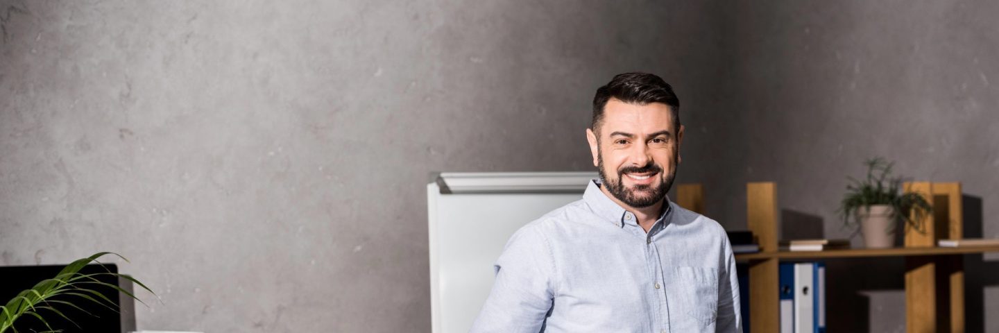 smiling businessman leaning on table in office and looking at camera
