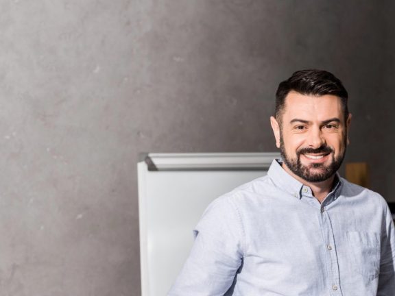 smiling businessman leaning on table in office and looking at camera