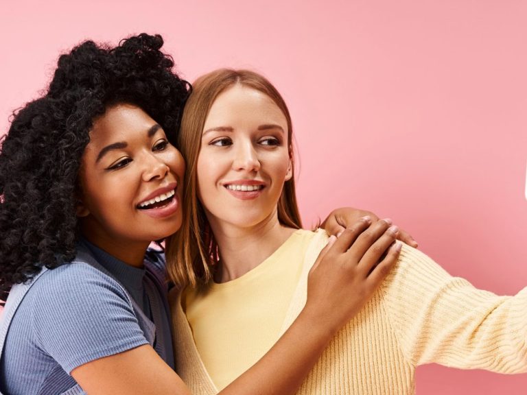 Two attractive diverse women in cozy casual attire taking a selfie with a cell phone.