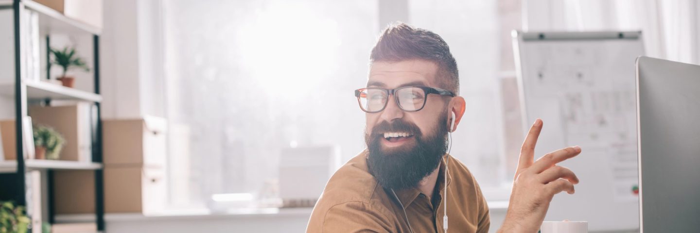 smiling bearded adult businessman in earphones sitting and working at computer desk in office