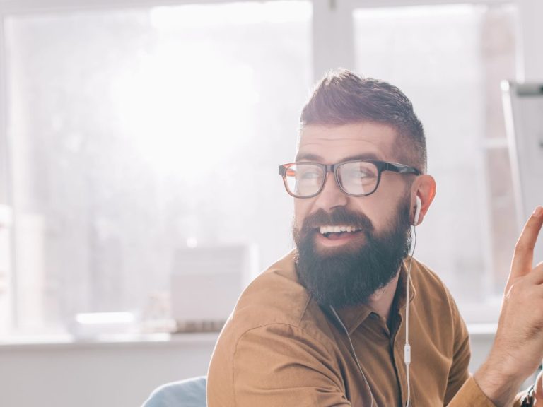 smiling bearded adult businessman in earphones sitting and working at computer desk in office