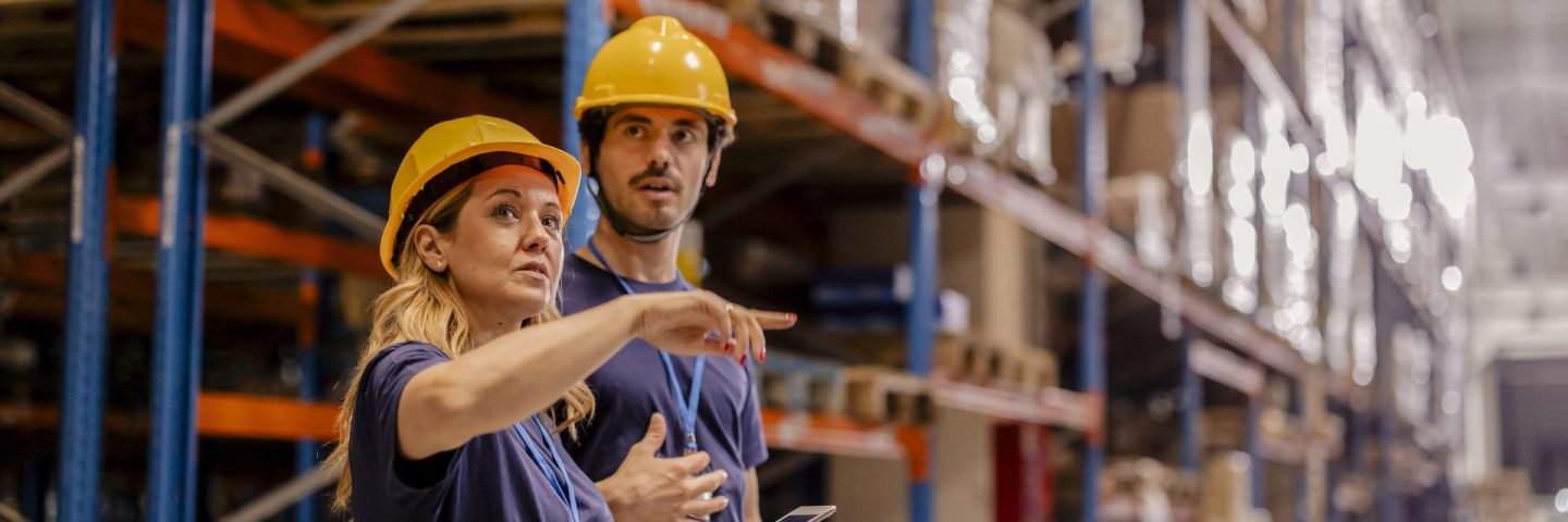 Two warehouse workers in safety gear discussing operations and reviewing inventory on a tablet in a warehouse aisle.