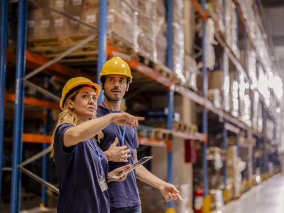 Two warehouse workers in safety gear discussing operations and reviewing inventory on a tablet in a warehouse aisle.