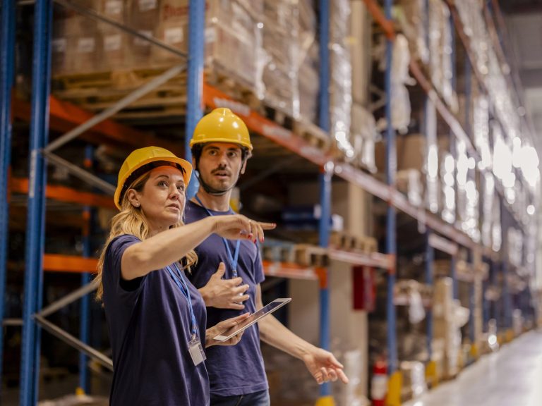 Two warehouse workers in safety gear discussing operations and reviewing inventory on a tablet in a warehouse aisle.